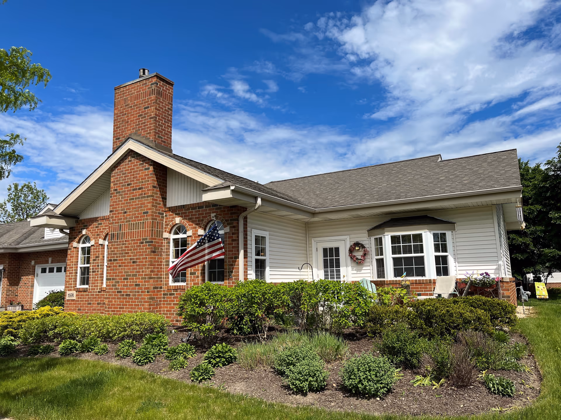 Exterior view of a single-story senior living facility building with a brick chimney, white siding, and a well-maintained garden with shrubs and flowers. An American flag is mounted near the windows, and the sky is partly cloudy with blue patches.