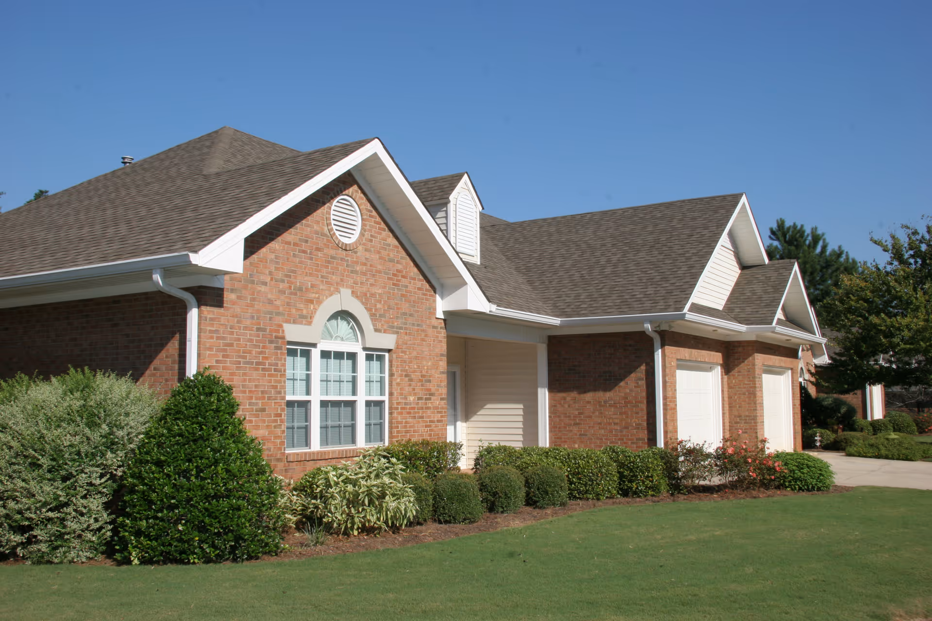Exterior view of a brick building with white trim, multiple windows, and a well-maintained lawn with shrubs and bushes under a clear blue sky.