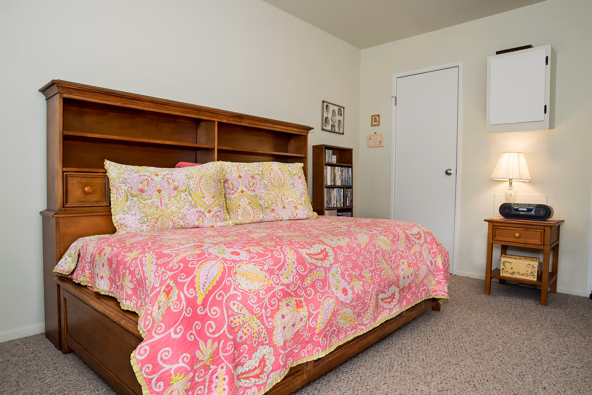 A bedroom with a wooden bed frame featuring built-in shelves, a pink and yellow patterned quilt and matching pillows. Next to the bed is a wooden nightstand with a lamp and a radio. In the background, there is a white door, a small bookshelf filled with DVDs or books, and a wall-mounted cabinet.