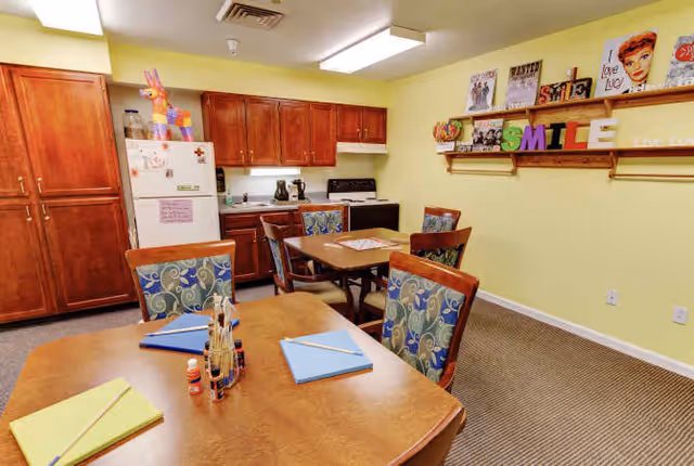Communal dining and kitchenette area with wooden tables and patterned chairs, a small kitchen with cabinets and refrigerator, and decorative shelves spelling 'SMILE'.