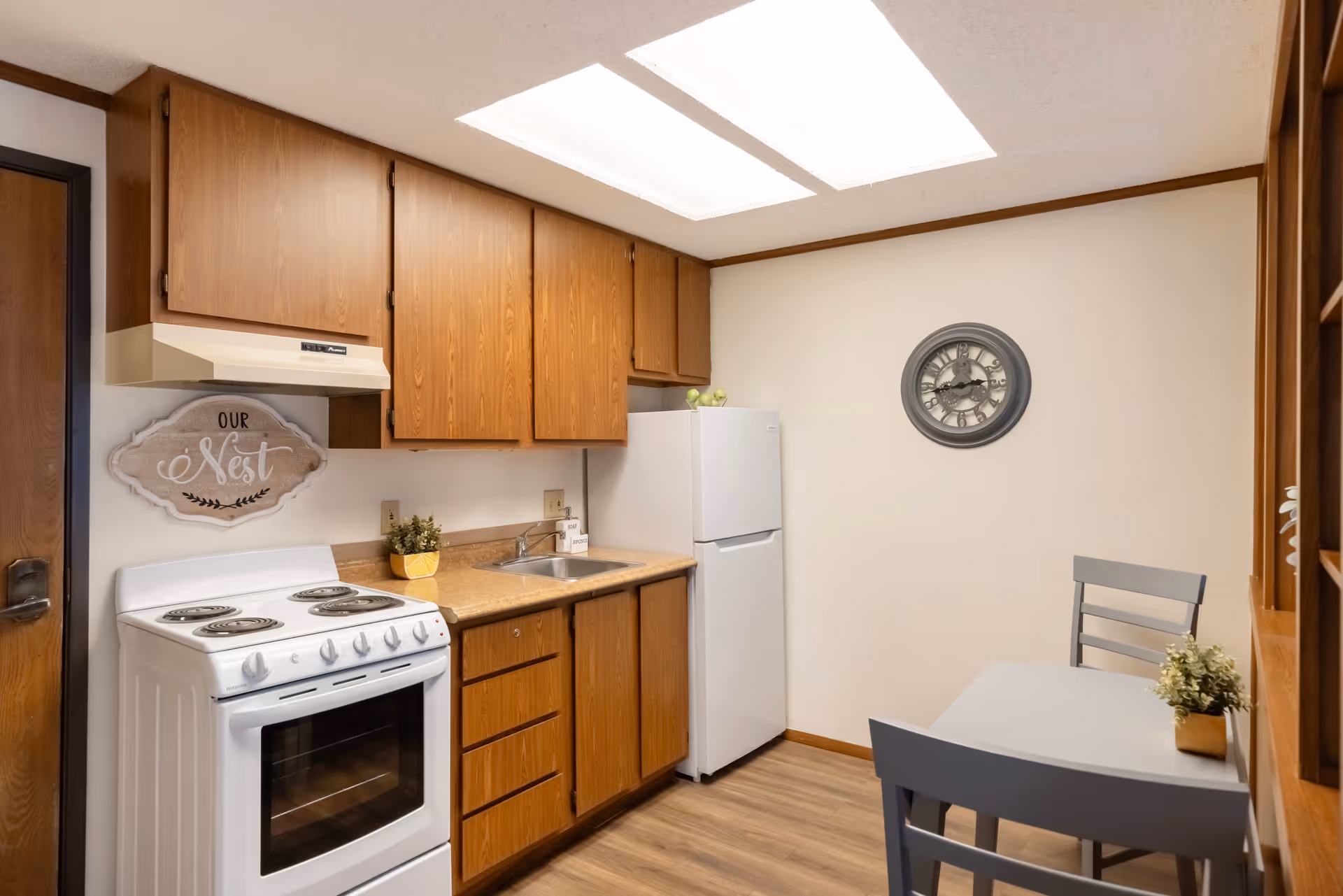 A small kitchen area with wooden cabinets, a white electric stove, a white refrigerator, a sink, and a small dining table with two chairs. There is a decorative sign on the wall that says 'Our Nest' and a round wall clock. The floor is wood and the ceiling has a rectangular light fixture.