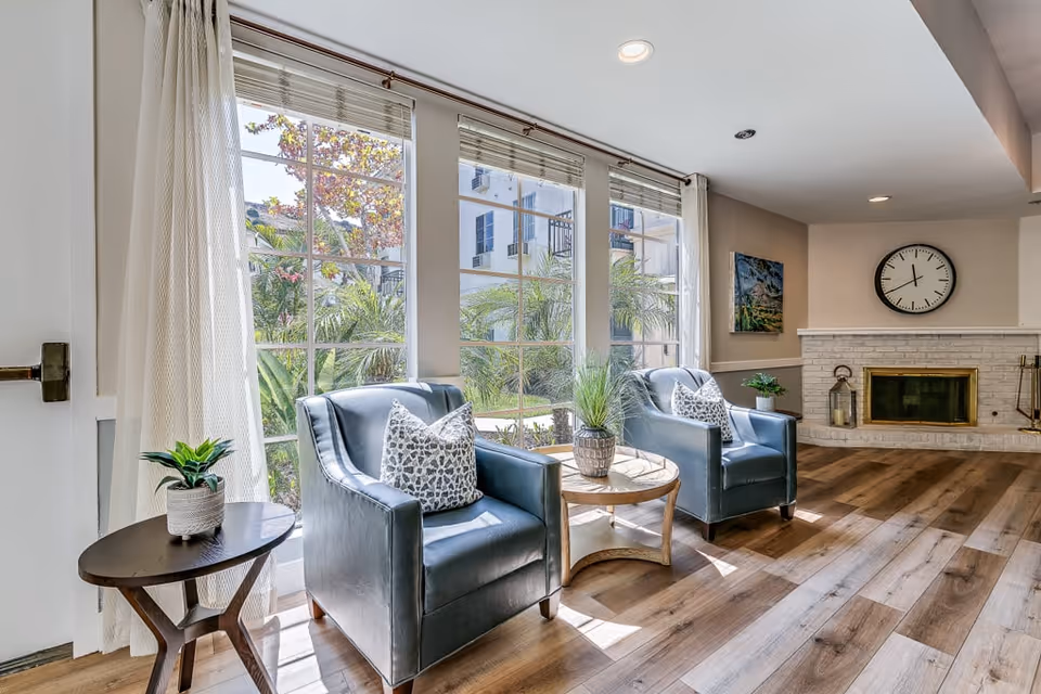 A bright and cozy living room area with two blue armchairs each with a patterned pillow, a round wooden coffee table with a plant, large windows with white curtains letting in natural light, a white brick fireplace with a clock above it, and wooden flooring.