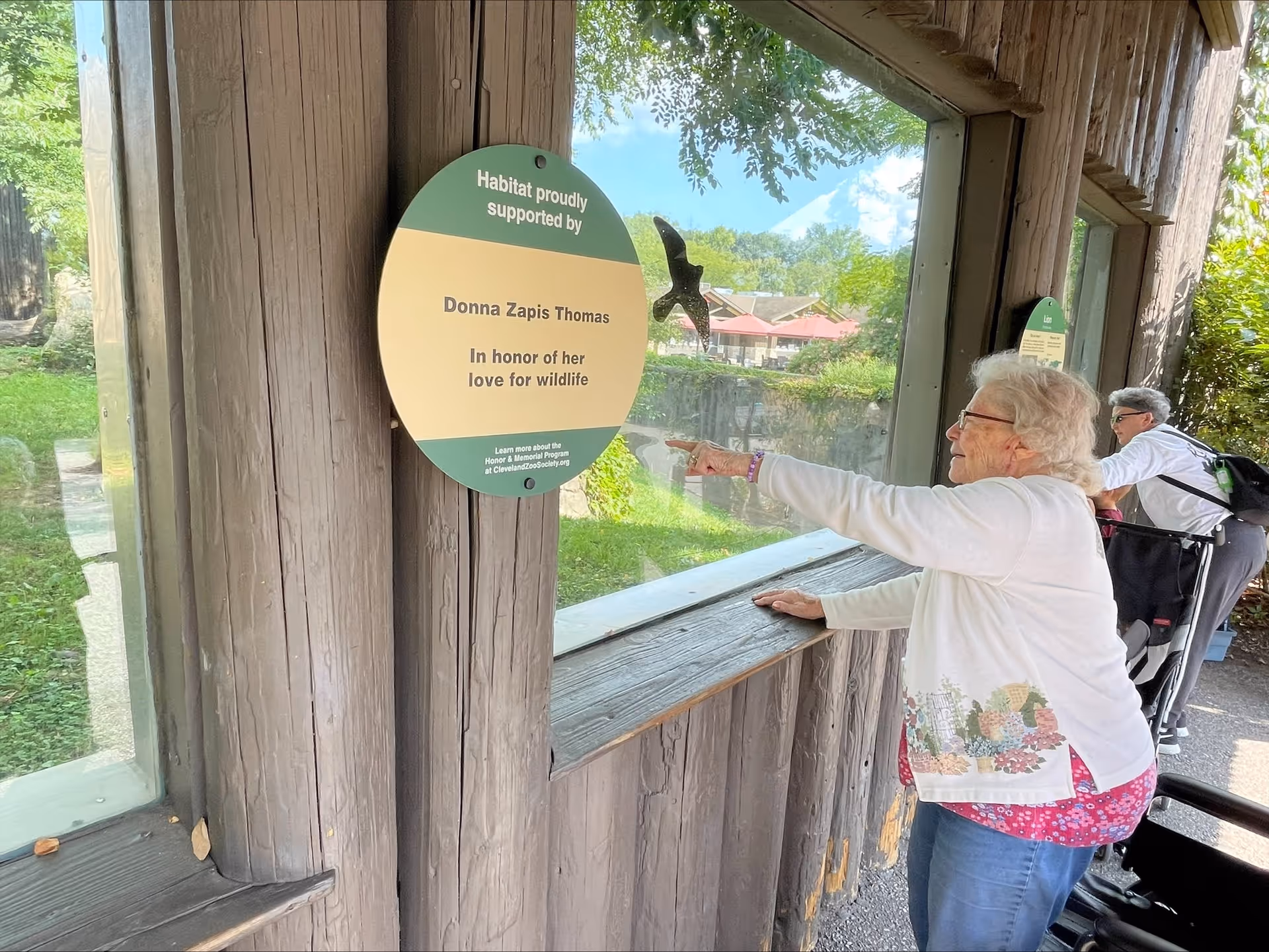 An elderly woman in a white sweater with floral patterns points at a bird inside a glass enclosure at a zoo or wildlife habitat. Another person in a wheelchair is nearby, also looking at the bird. A sign on the wooden wall reads, 'Habitat proudly supported by Donna Zapis Thomas In honor of her love for wildlife.'