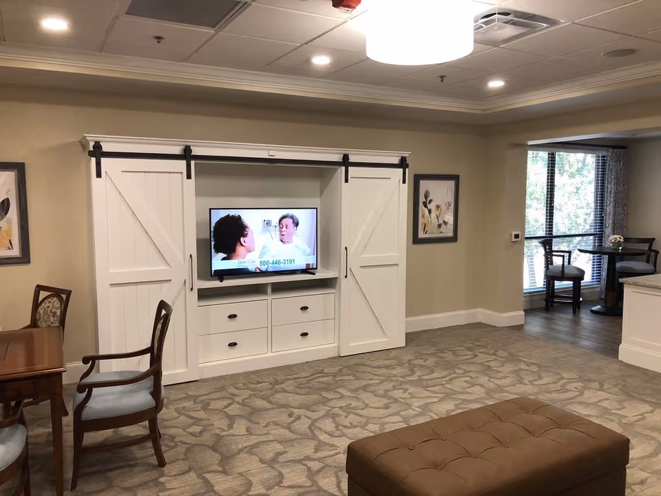 A cozy living area in a senior living facility featuring a TV set inside a white cabinet with sliding barn doors. The TV is on, showing two people talking. There is a wooden dining table with chairs on the left side, a brown cushioned ottoman in the foreground, and a small seating area with two chairs and a round table near a window with blinds in the background. The room has beige walls, carpeted flooring, and recessed ceiling lights.