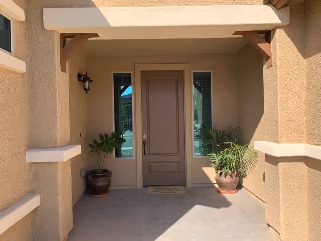 Front entrance of a building with a brown door centered between two narrow windows. There are two potted plants on either side of the door and a small outdoor light fixture mounted on the left wall. The walls are textured and painted beige, and the entrance area is shaded by an overhang supported by wooden brackets.