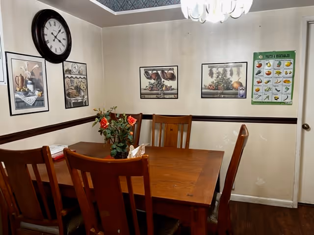 Wood dining table with six chairs, a floral centerpiece, a wall clock, framed prints and a chandelier in a small dining room.
