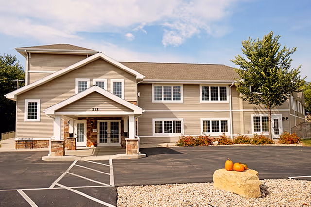Exterior view of a two-story beige assisted living facility building with multiple windows, a covered entrance, a parking lot, and a landscaped area with a large rock and three pumpkins. A tree is visible on the right side under a partly cloudy sky.