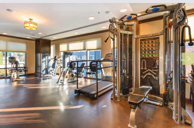 Bright and spacious fitness room with various exercise equipment including treadmills, stationary bikes, and a multi-functional weight machine. Large windows allow natural light to fill the room.