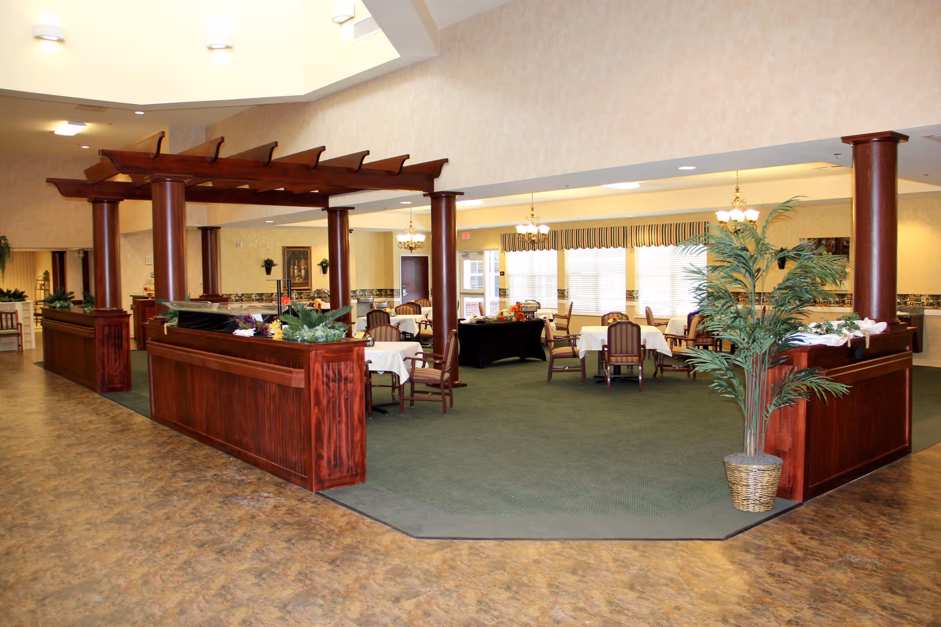 Interior view of a senior living facility dining area with multiple tables covered in white tablecloths and surrounded by wooden chairs with striped cushions. The space features a green carpeted floor section bordered by a wooden structure with columns and decorative plants. Large windows with blinds and striped valances allow natural light to fill the room, and chandeliers hang from the ceiling providing additional lighting.