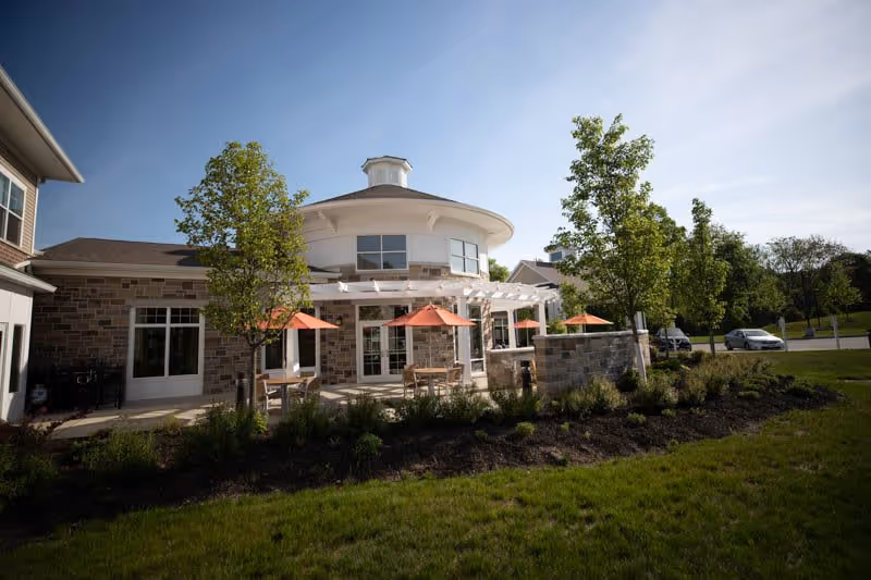 Exterior view of Vista Springs Ravinia Estate showing a building with stone and brick facade, a round turret-like structure with windows, outdoor patio area with tables and orange umbrellas, surrounded by green grass, trees, and shrubs under a clear blue sky.