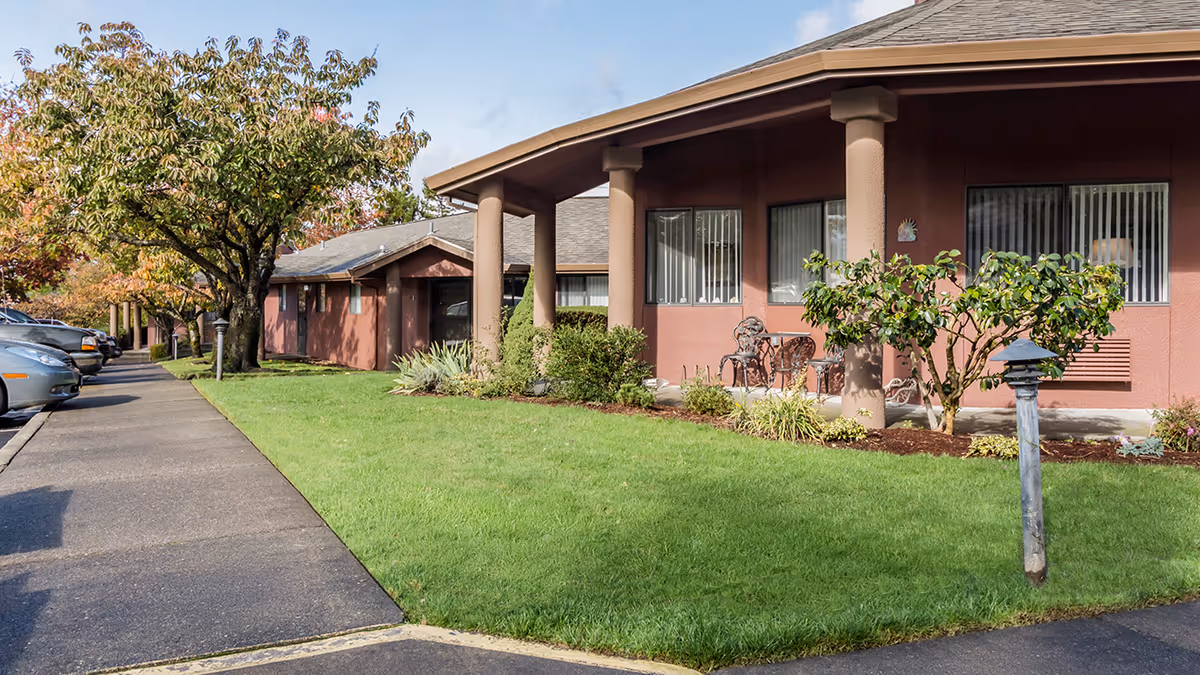 Exterior view of a single-story building at Capital Place Retirement Community with a covered porch supported by columns, outdoor seating, green lawn, trees, and a sidewalk adjacent to a parking area with several cars.