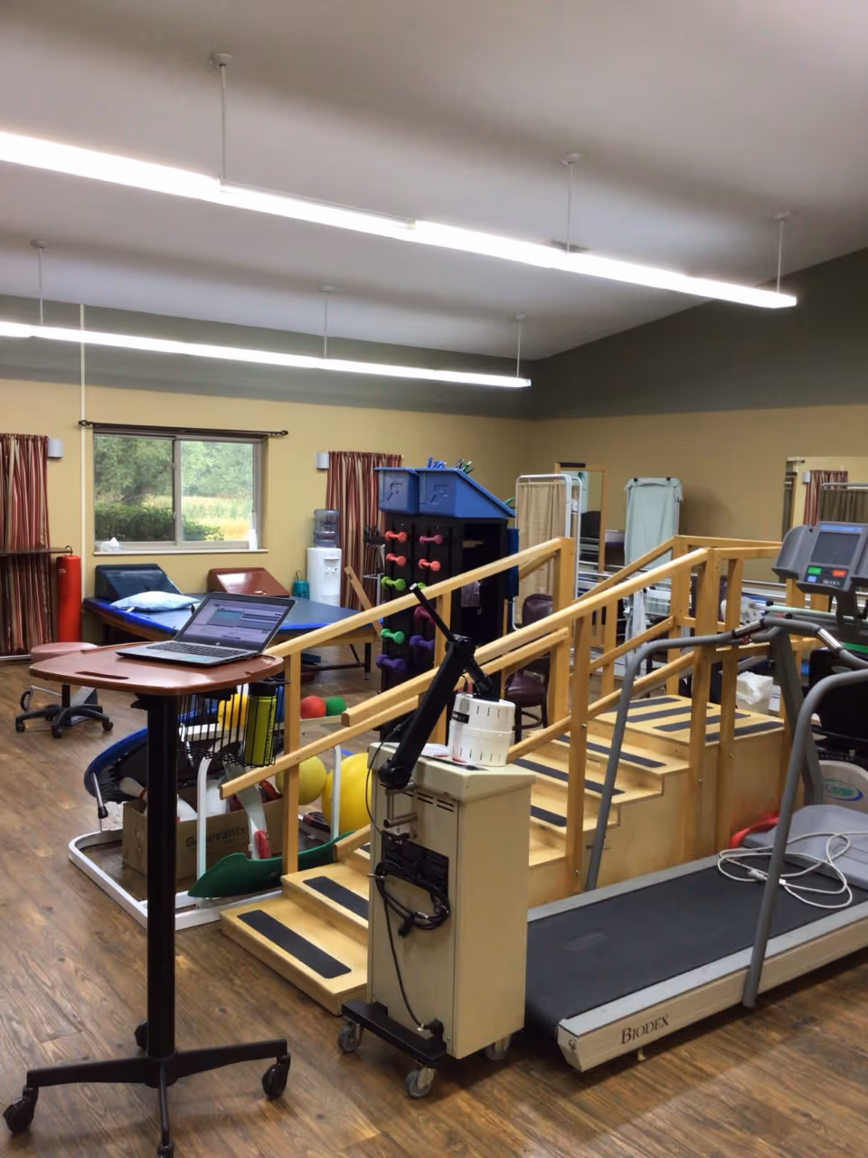 Physical therapy room with wooden stairs and handrails for rehabilitation exercises, a treadmill, a table with a laptop, exercise balls, dumbbells on a rack, and therapy tables near windows with curtains.