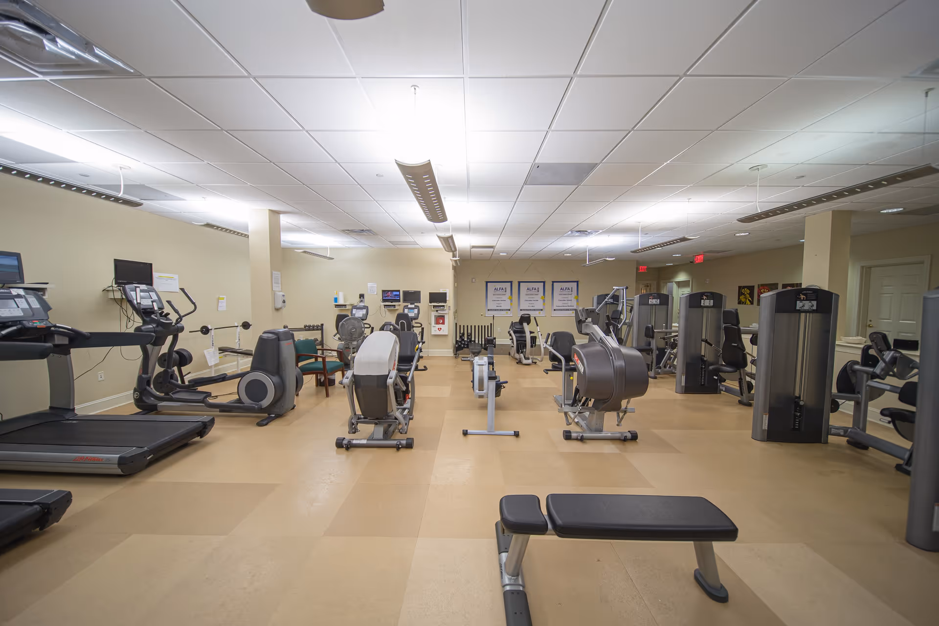 A spacious indoor fitness room with various exercise equipment including treadmills, stationary bikes, rowing machines, weight machines, and benches. The room has beige walls, a tiled ceiling with fluorescent lights, and a tan floor. There are posters on the far wall and a few chairs along the left side.