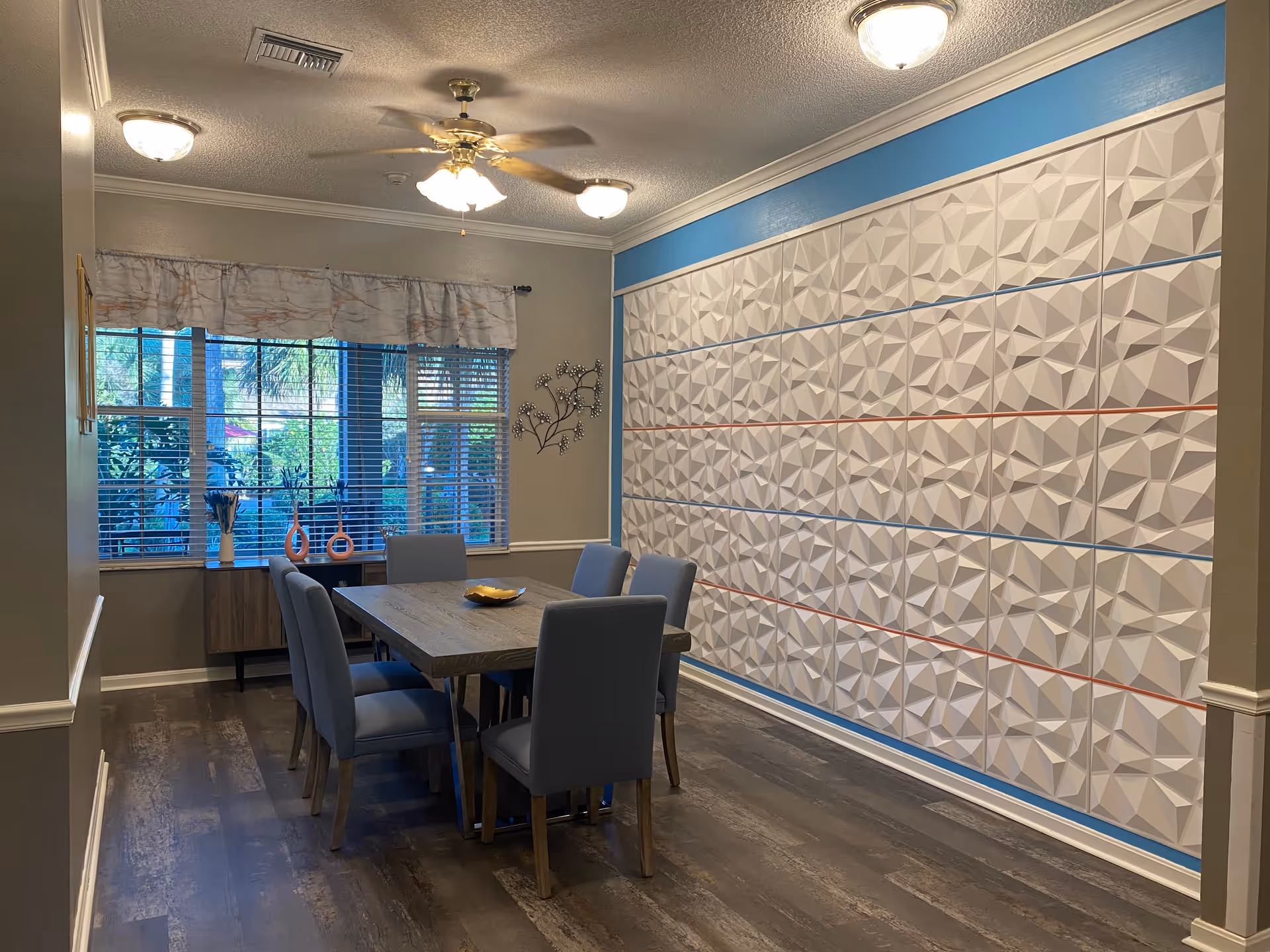 A dining room with a wooden table surrounded by six gray upholstered chairs. The room features a large window with blinds and a valance, a decorative wall with geometric 3D panels in white with blue and red lines, and a ceiling fan with lights. There is a small cabinet under the window with decorative vases on top.