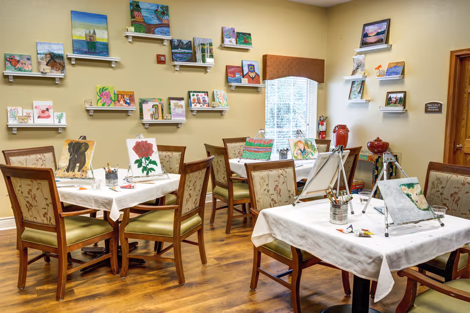 A bright art room with several tables covered in white tablecloths, each set up with easels, paintbrushes, and paintings in progress. The walls are decorated with multiple colorful paintings displayed on small white shelves. The room has wooden floors and upholstered chairs with floral patterns. A window with blinds and a brown valance lets in natural light.