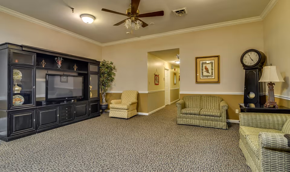 A senior living facility common area with patterned carpet, beige walls with white trim, and a ceiling fan with lights. The room features a large black entertainment center with a flat-screen TV, a potted plant, a beige armchair, two green patterned sofas, a tall black grandfather clock, a table lamp, and framed artwork on the walls. A hallway is visible in the background.