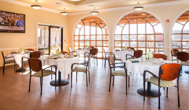 Sunlit dining room with round tables set for meals and chairs arranged near large arched windows.