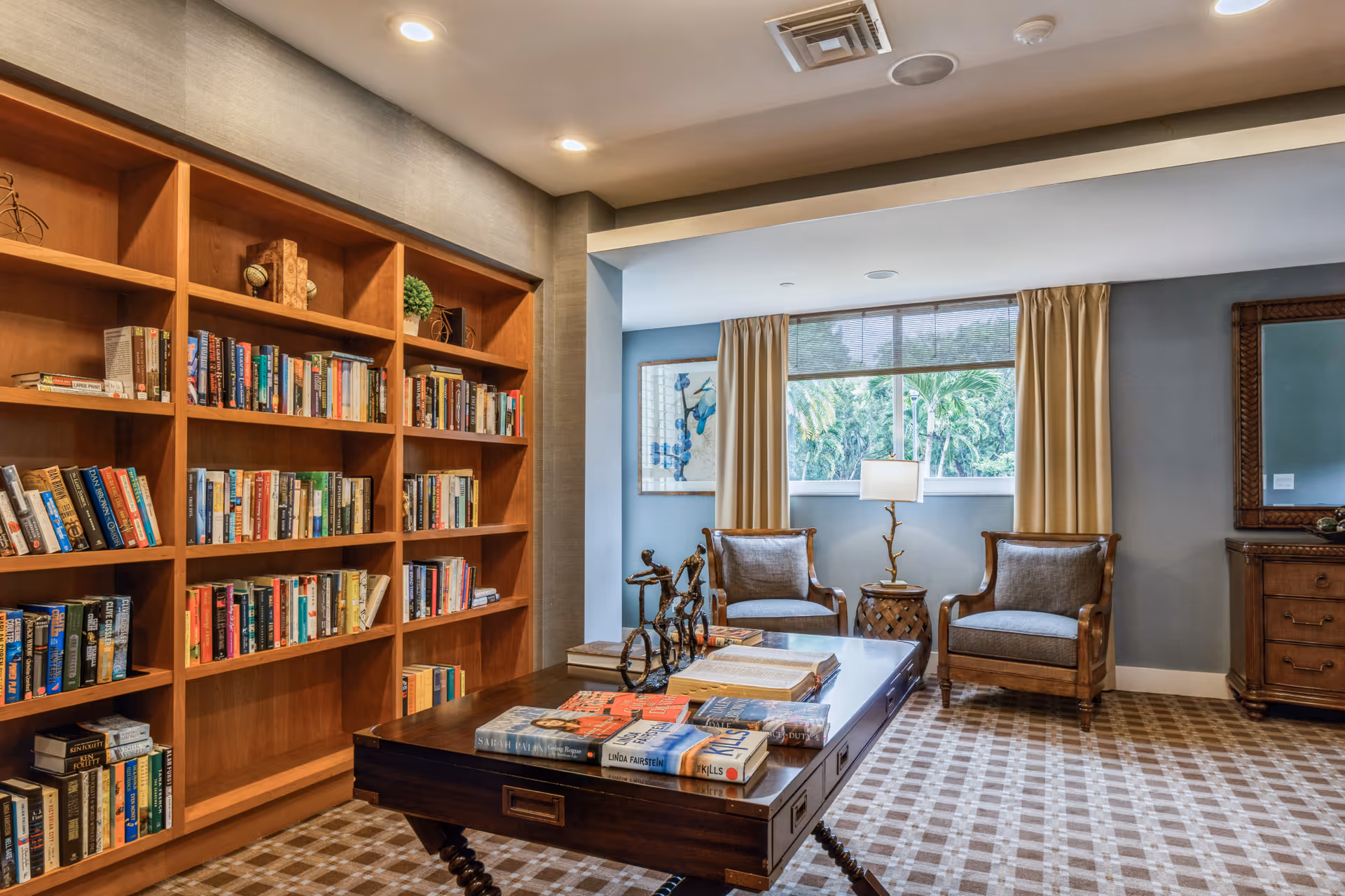 A cozy reading room with a large wooden bookshelf filled with books on the left, a wooden table in the center with books and decorative items on it, two cushioned armchairs near a window with beige curtains, a table lamp, and a wooden dresser with a mirror on the right. The room has a patterned carpet and soft lighting.