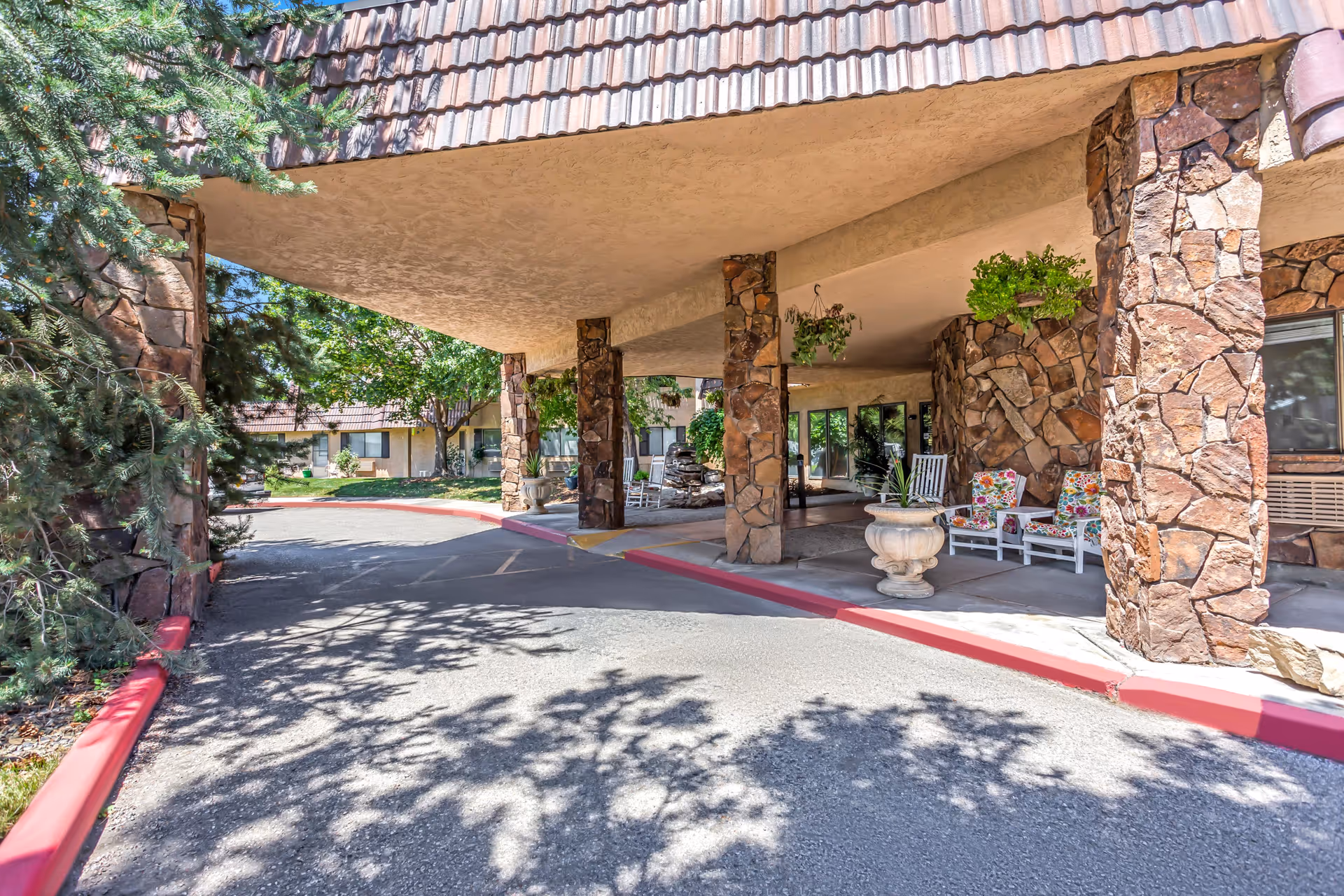 Covered entrance area of a senior living community with stone pillars, hanging plants, and white chairs with floral cushions. Trees and greenery surround the driveway leading to the entrance.