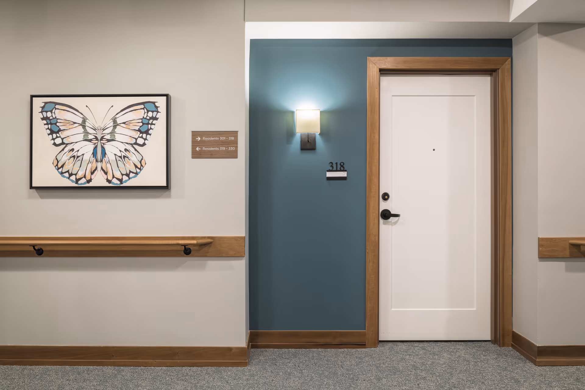 A hallway in a senior living facility with a white door numbered 318 framed in wood. The wall around the door is painted teal with a wall-mounted light above the number. To the left, there is a large framed butterfly artwork and a wooden handrail running along the wall. A small sign indicates directions to residents' rooms 301-318 and 319-330.