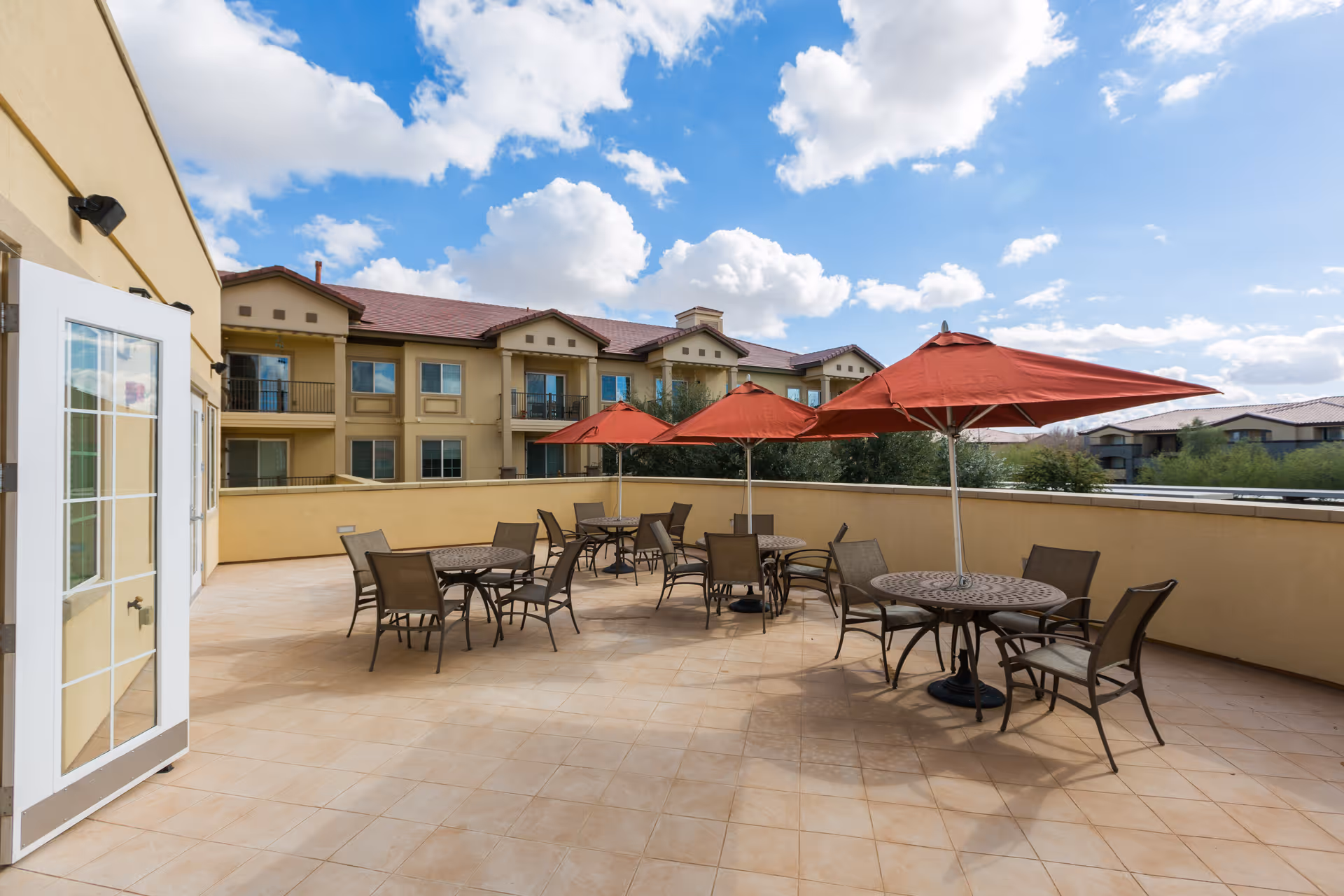 Outdoor patio area with several round tables and chairs, each table shaded by a large red umbrella. The patio is tiled and surrounded by a low wall, with a multi-story building in the background under a partly cloudy blue sky.