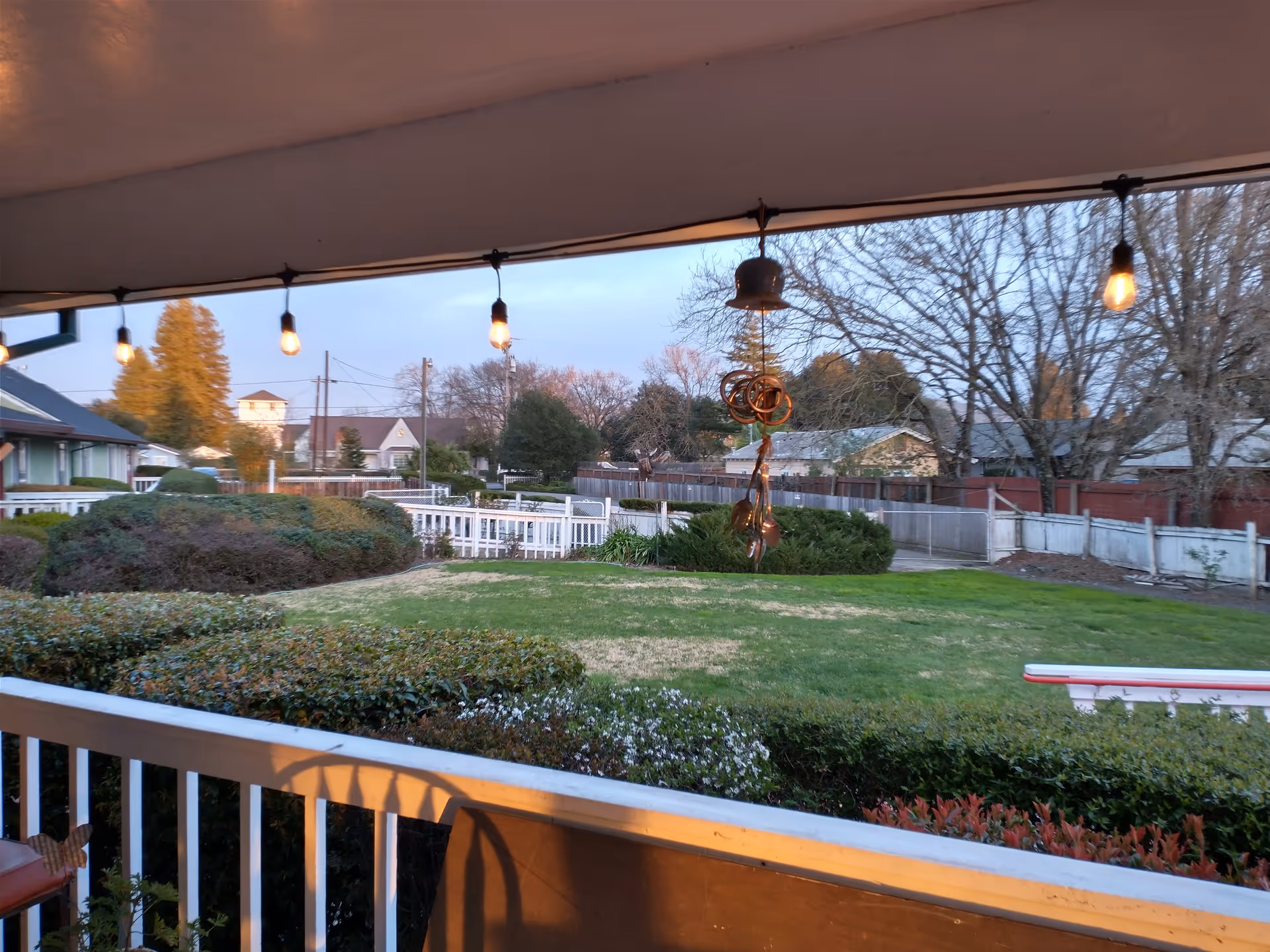 View from a covered porch with hanging string lights and a wind chime, overlooking a green lawn, bushes, and trees with houses and a fence in the background during early evening.