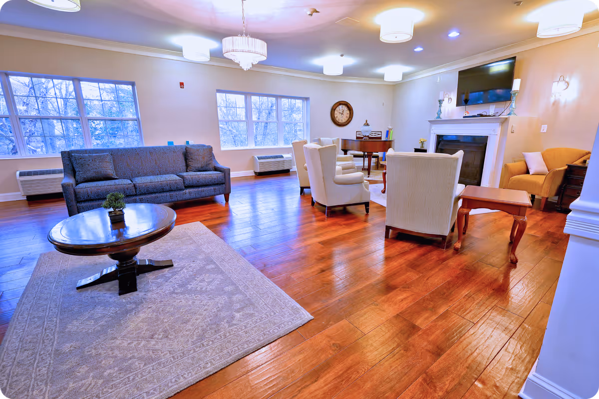 A spacious and well-lit living room with wooden flooring, a gray sofa, several beige armchairs arranged around a coffee table, a fireplace with a mounted TV above it, a wall clock, and large windows letting in natural light.