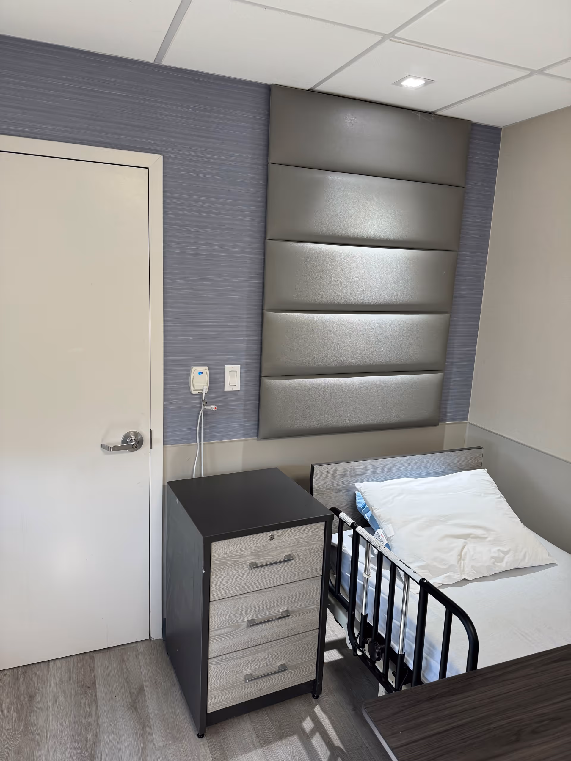 A small nursing facility room with a single bed featuring a white pillow and a black safety rail. Next to the bed is a three-drawer nightstand with a black top and light wood drawer fronts. The wall behind the bed has padded gray panels and a light switch with a call button. The floor is light wood, and there is a closed white door to the left.