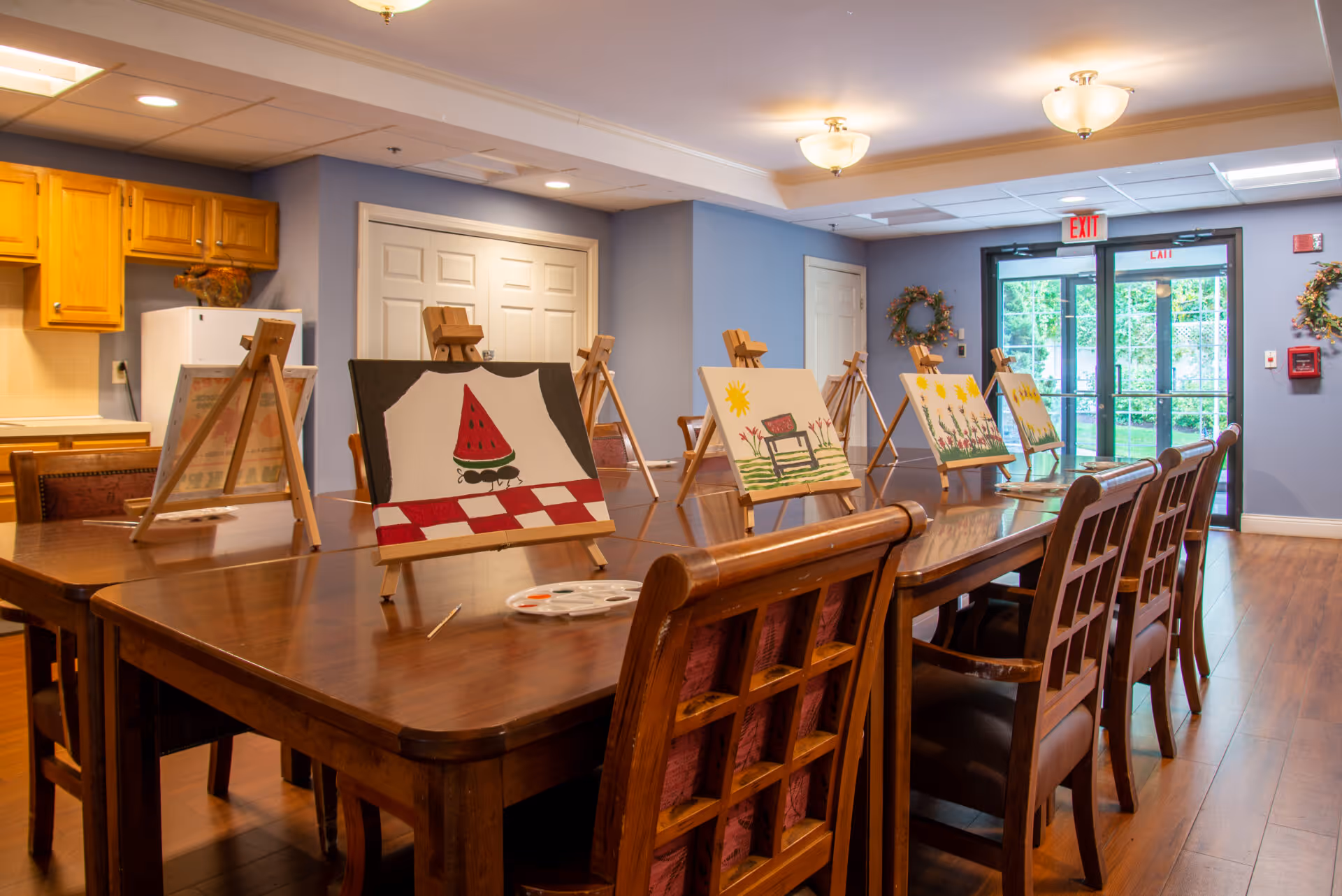 Activity room with a long wooden table and easels displaying paintings, a kitchenette to the left and glass exit doors at the back.