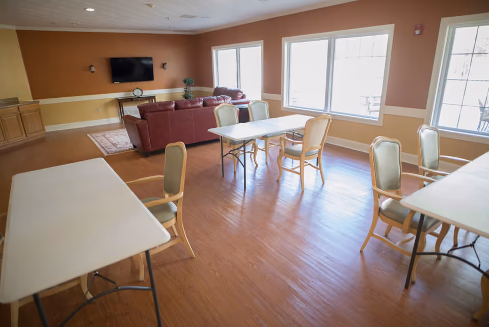A spacious room with several tables and chairs arranged for seating, a red leather couch set near a wall-mounted TV, large windows letting in natural light, and wooden flooring. The walls are painted in warm tones of orange and beige.