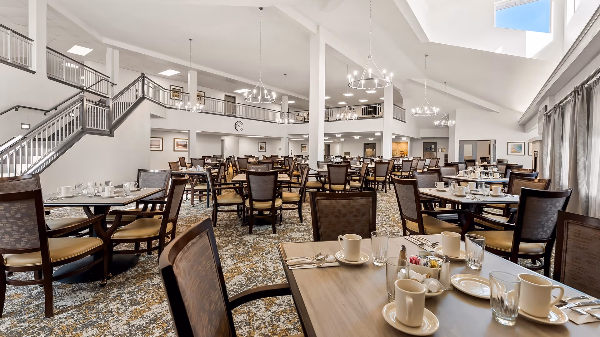 Spacious dining room with multiple wooden tables and chairs arranged neatly. Each table is set with cups, glasses, plates, and silverware. The room features high ceilings with modern chandeliers, large windows with curtains, and a staircase leading to an upper level with a railing. The carpet has a patterned design, and the walls are decorated with framed pictures.