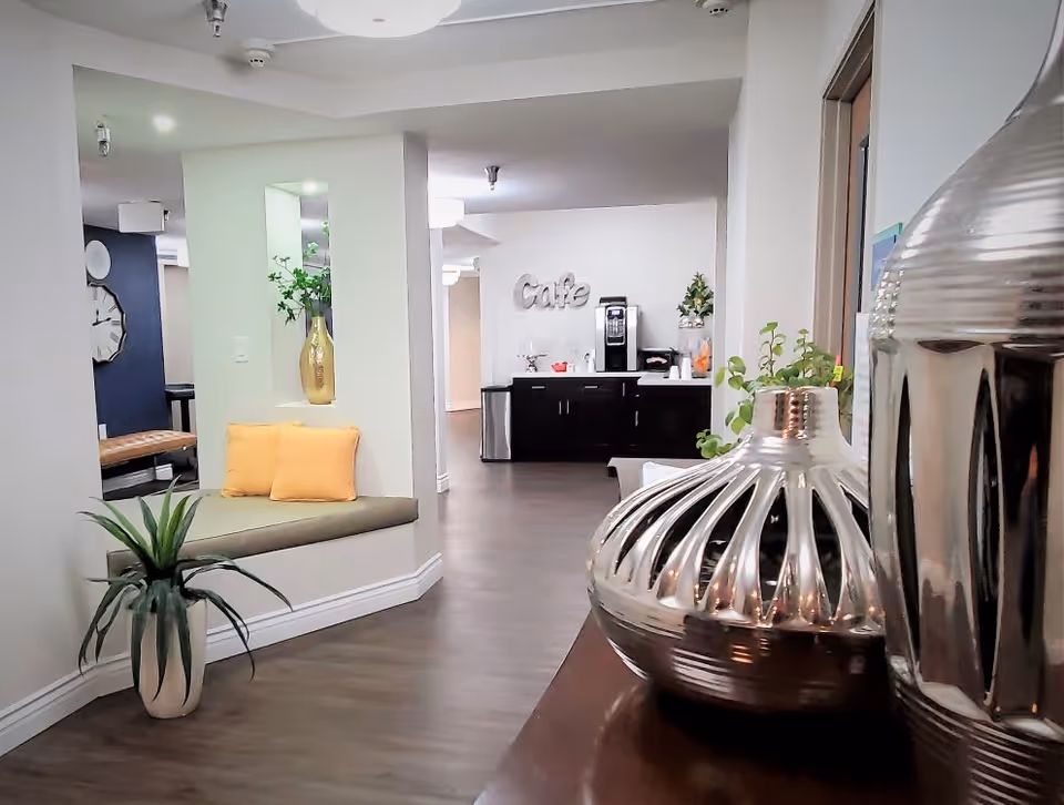 Interior view of a senior living facility hallway with a built-in bench featuring two yellow cushions, a potted plant on the floor, decorative silver vases on a wooden surface, and a cafe area with a coffee machine and the word 'Cafe' on the wall in the background.