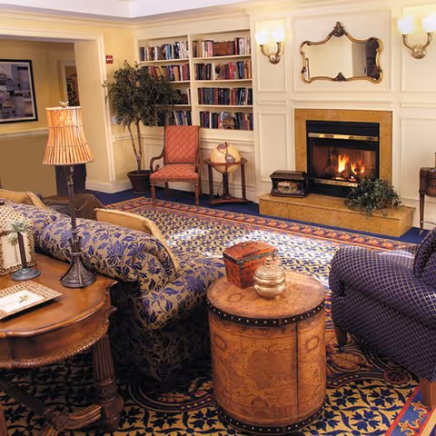 Cozy living room area with a lit fireplace, patterned sofas and armchairs, a wooden side table with a lamp, a round wooden chest, a bookshelf filled with books, and a potted plant in the corner.