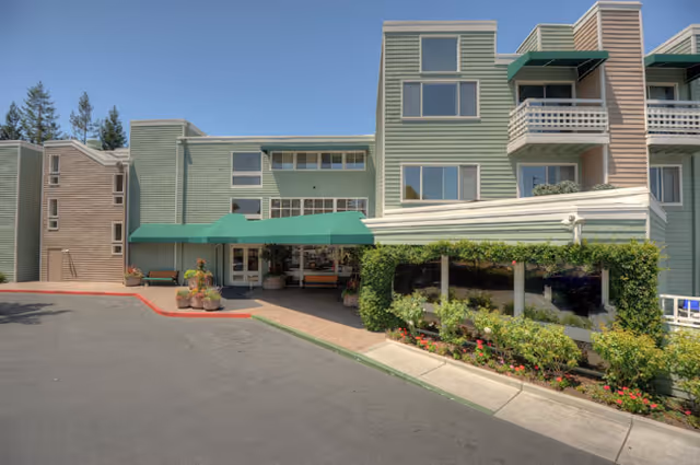 Exterior view of a multi-story senior living facility building with green siding and multiple windows. The entrance has a green awning, and there are plants and flowers along the sidewalk near the building. The sky is clear and blue.