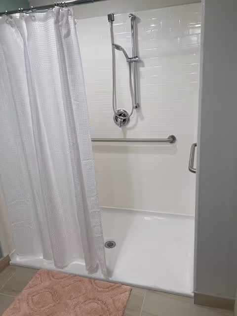 A clean, accessible shower area with white tiled walls, a silver adjustable showerhead, a horizontal grab bar, and a white shower curtain partially drawn. A pink bath mat is placed on the tiled floor outside the shower.