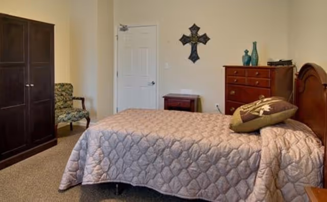 A bedroom with a single bed covered with a quilted beige bedspread and a decorative pillow. There is a wooden dresser with vases and other items on top, a small wooden nightstand, a dark wooden wardrobe, and a patterned armchair. A decorative cross hangs on the wall above the nightstand.