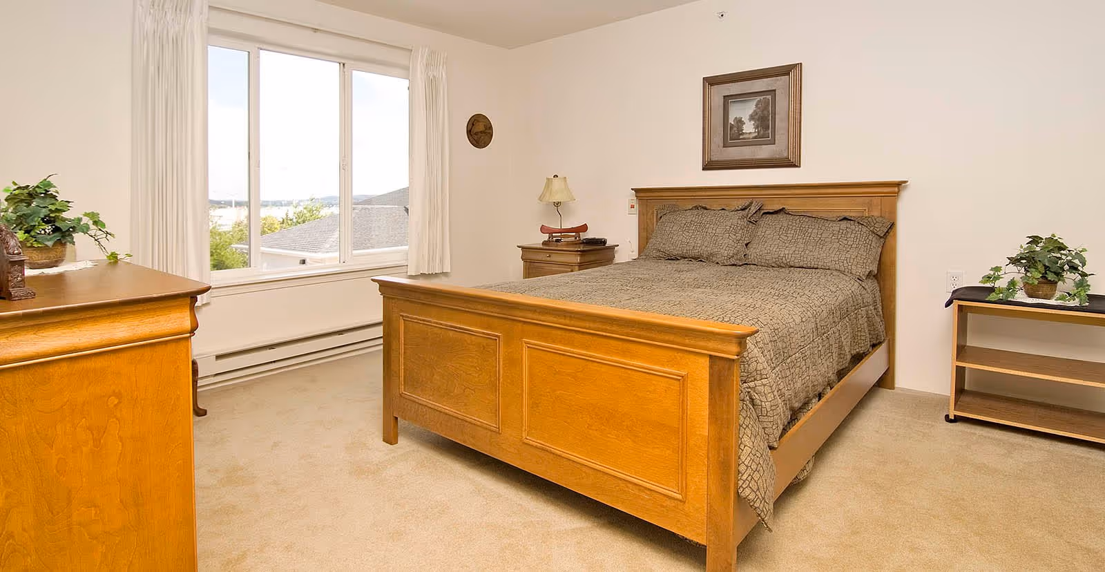 A bedroom with a wooden bed frame and matching furniture including a dresser and nightstand. The bed is made with a patterned brown bedspread and pillows. There is a window with white curtains letting in natural light, a framed picture on the wall above the bed, and two potted plants on the dresser and a small table.