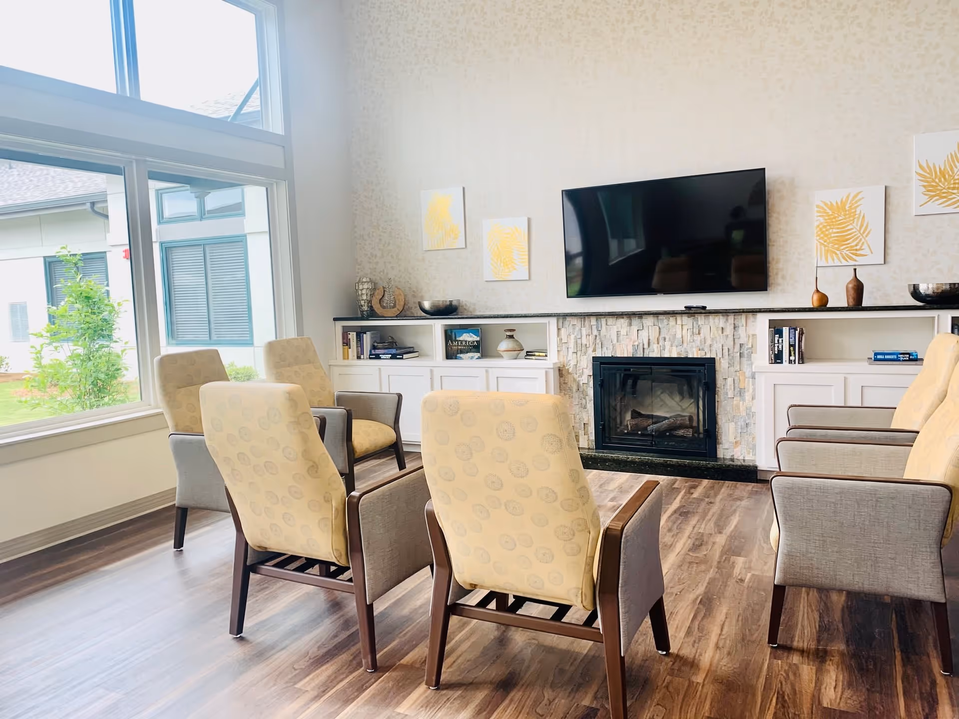 A bright and cozy living room with six upholstered armchairs arranged in a semi-circle facing a wall-mounted flat-screen TV above a stone fireplace. The room has large windows letting in natural light, wooden flooring, built-in white cabinets with books and decorative items, and three framed artworks with yellow leaf designs on the wall.