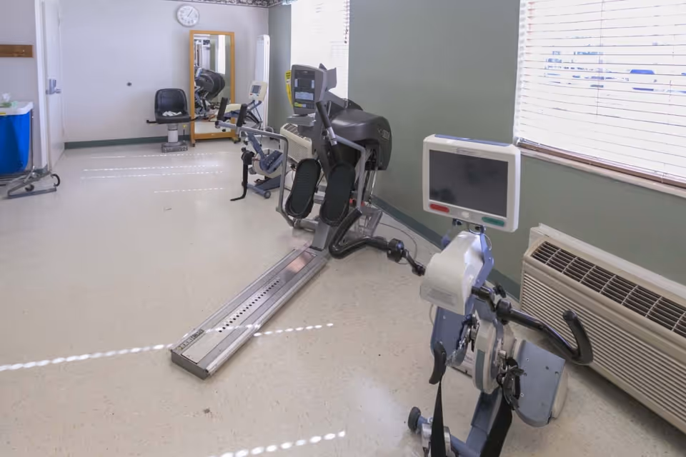 A rehabilitation room with exercise equipment including a recumbent bike and a rowing machine, a chair, a mirror on the wall, a clock, and a window with blinds letting in natural light.