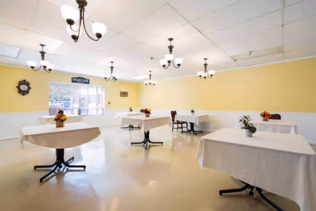 A bright dining room with several tables covered in white tablecloths, each decorated with small flower arrangements. The walls are painted yellow with white wainscoting, and multiple ceiling light fixtures hang from a white tiled ceiling. A window with blinds and a welcome sign is visible on one wall.