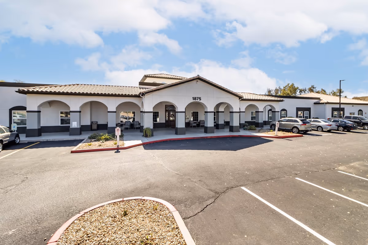 Front exterior view of a single-story building with white walls and a tiled roof, featuring multiple arches along the entrance. Several cars are parked in the parking lot in front of the building under a partly cloudy sky.