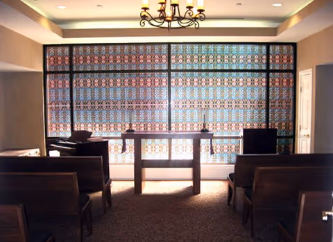 Interior room with rows of wooden benches facing a small wooden table and a piano near a large window covered with decorative patterned glass panels. A chandelier hangs from the ceiling.