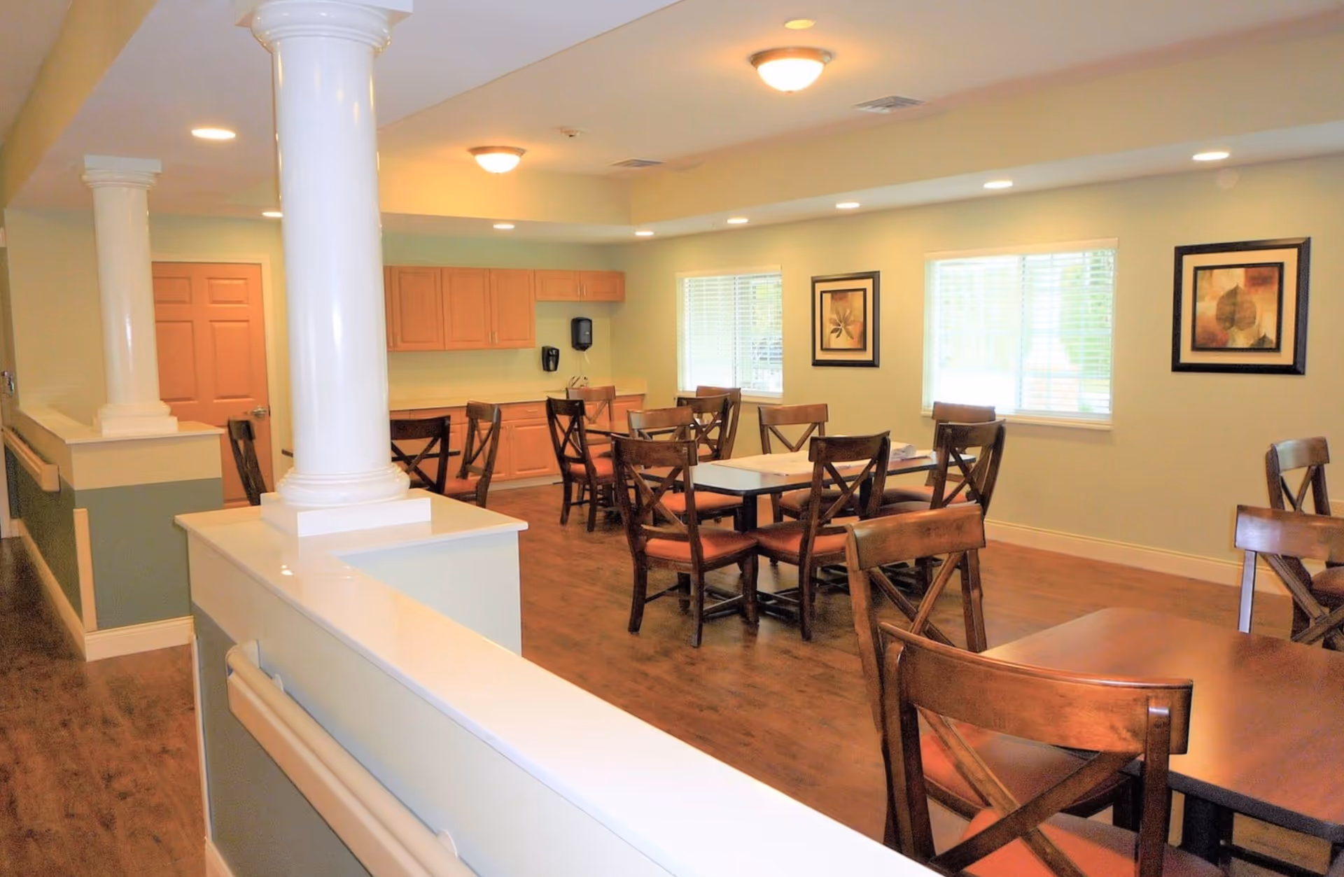 A bright dining room with wooden tables and chairs arranged neatly. The room features light green walls, two windows with blinds, framed artwork on the walls, and a ceiling with recessed lighting and a central light fixture. There are white decorative columns and a wooden floor.