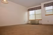 Empty room with beige carpet, white walls, two windows with blinds and valances, and a wall-mounted air conditioning unit below one window.