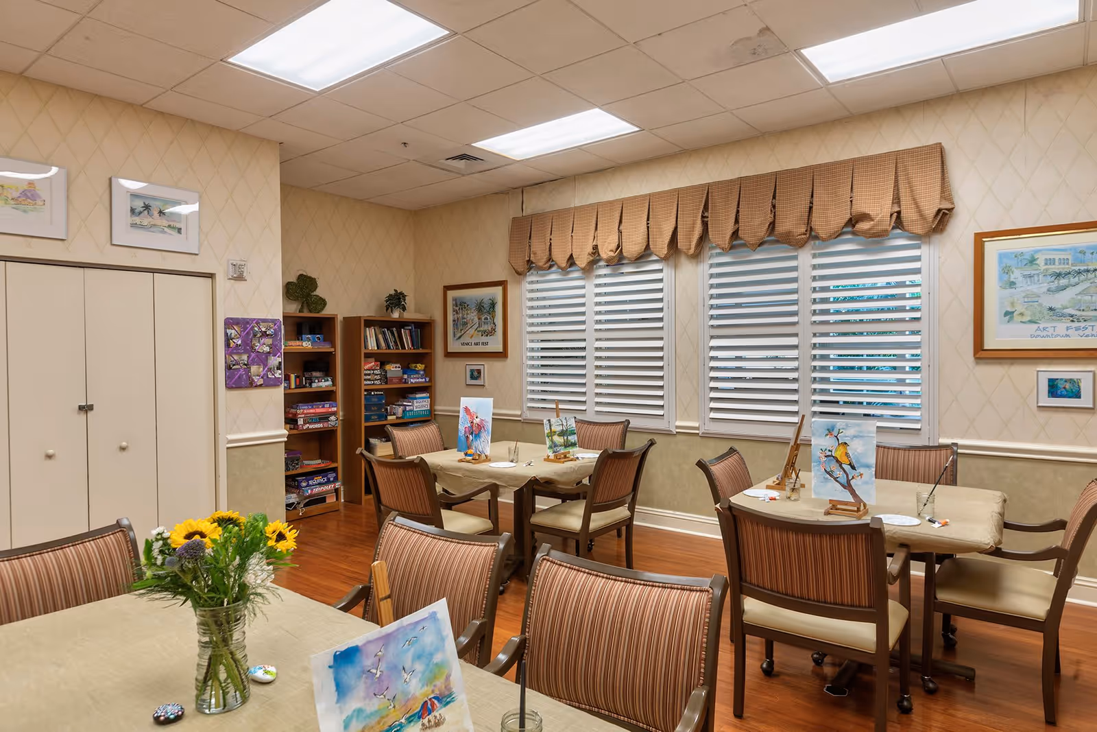 A well-lit activity room with several tables and chairs arranged for painting. Each table has easels with paintings of birds and nature scenes. There is a vase with sunflowers on one table. The room has beige walls with framed artwork, a bookshelf filled with board games and books, and windows with white blinds and brown valances.