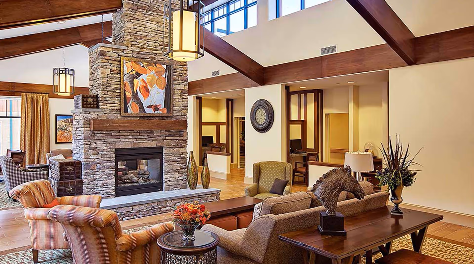 Spacious senior living common room with a stone double-sided fireplace, upholstered chairs and sofas, wooden beams, and decorative accents.