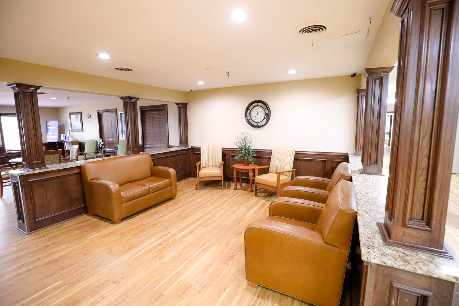 A spacious seating area in a senior living facility with wooden flooring, brown leather couches, two striped armchairs, a small wooden table with a plant, and a wall clock. The room features wooden columns and trim, with a granite countertop along one side.