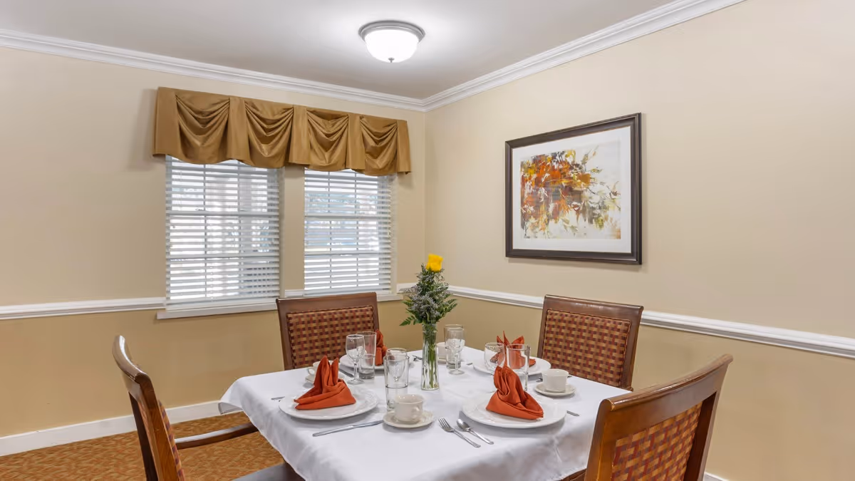 A dining room with a table set for four featuring white linens and folded red napkins, chairs, a vase with flowers, and framed artwork on beige walls.