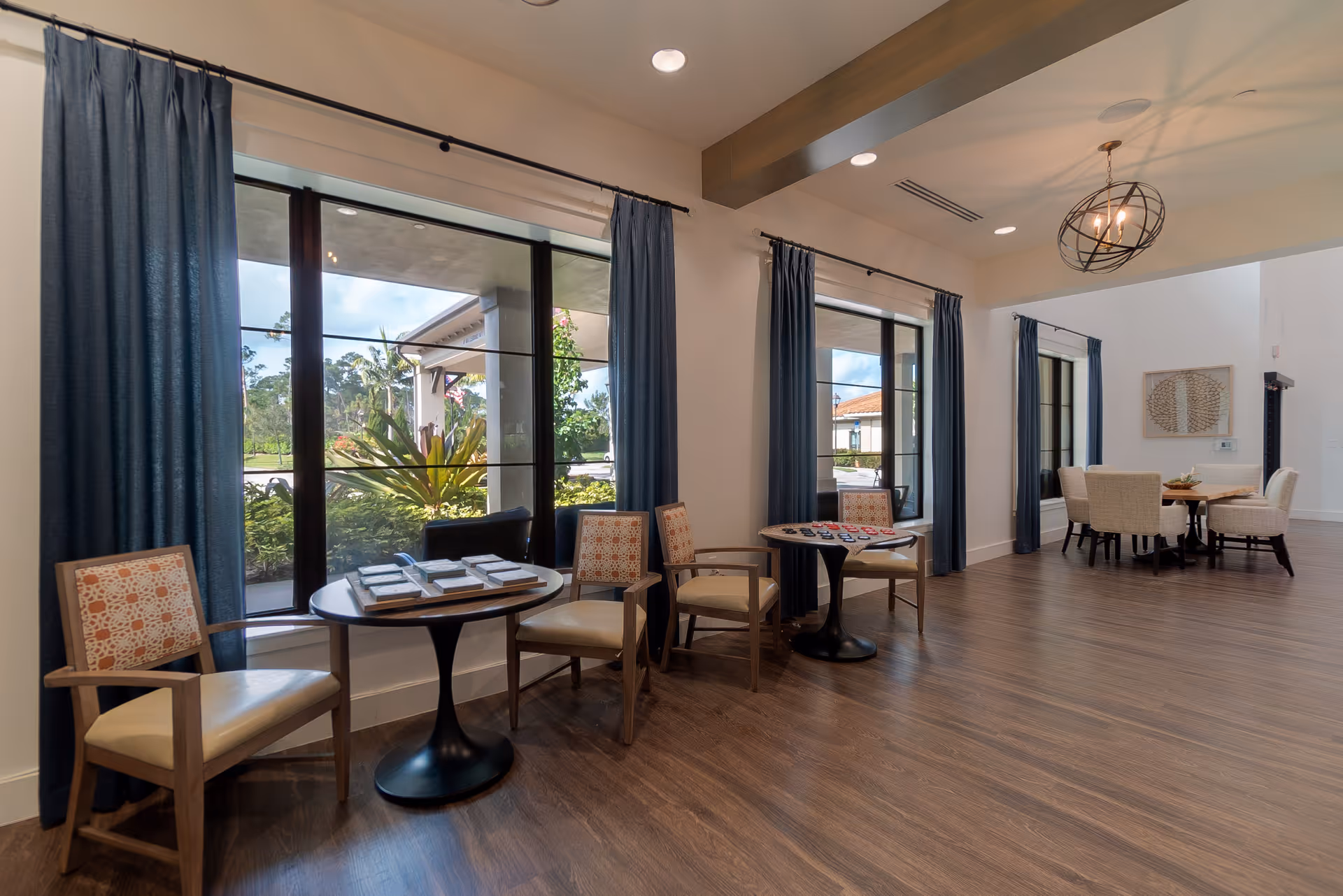 A spacious, well-lit common area with large windows covered by blue curtains. The room features wooden flooring, several small round tables with chairs, and a modern chandelier hanging from the ceiling. Outside the windows, greenery and part of the building exterior are visible.