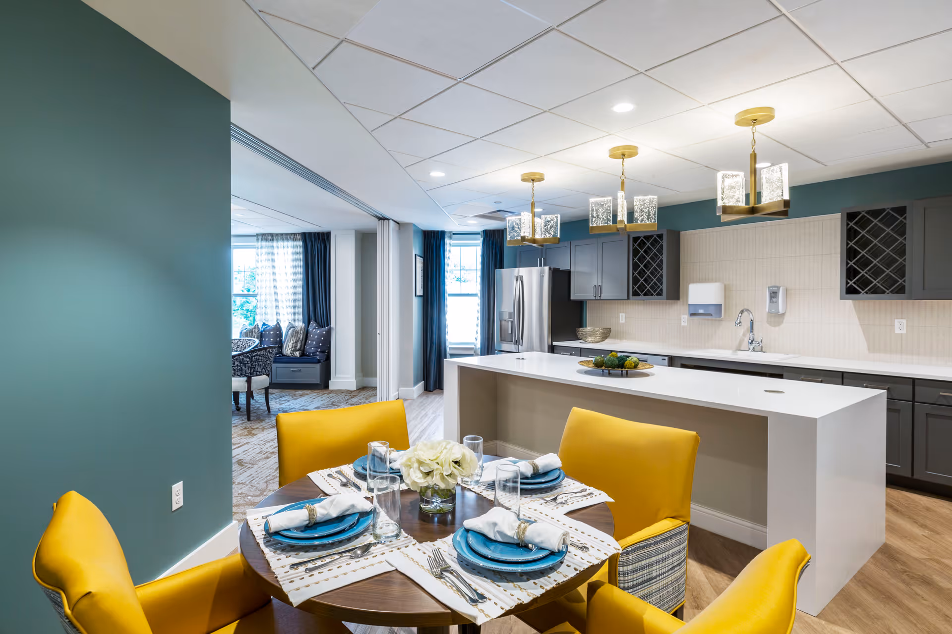 A modern kitchen and dining area in a senior living facility. The kitchen features a large white island with three pendant lights above it, dark gray cabinets, a stainless steel refrigerator, and a sink. In the foreground, there is a round wooden dining table set with blue plates, white napkins, silverware, and clear glasses, surrounded by four yellow upholstered chairs. The room has a green accent wall and large windows with blue curtains in the background.